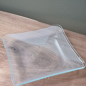 May include: A square glass plate featuring a series of concentric white lines that form a geometric pattern, radiating from the center toward the edges. The plate is transparent with smooth, rounded corners and a clear, polished finish throughout.