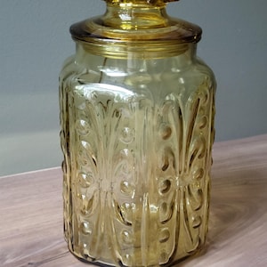 May include: A translucent amber-colored glass canister with a matching lid. The jar features a repeating embossed pattern of vertical lines and circular indentations along its exterior walls. The lid has a flat, circular knob handle on top.