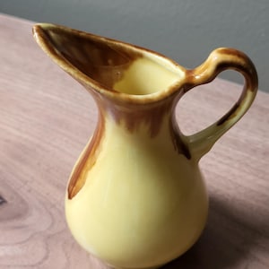 May include: A small ceramic pitcher featuring a pale yellow base color with glossy brown drip glaze accents around the rim, the handle, and the pouring spout. The vessel has a rounded body, a curved handle, and a pointed spout for pouring liquids.
