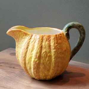 May include: A ceramic pitcher shaped like a yellow pumpkin with a textured, ribbed surface and a curved, dark green handle. The interior of the vessel is smooth and cream-colored, featuring a wide spout for pouring liquids.