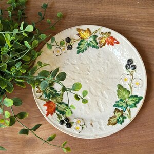May include: A round ceramic plate with a textured cream-colored surface, decorated with raised, hand-painted motifs of green leaves, autumn-colored foliage in orange and yellow, small white flowers, and clusters of dark berries along the rim.