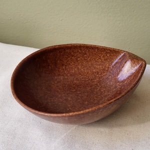 May include: A teardrop-shaped ceramic bowl with a speckled brown glaze. The bowl features a smooth, rounded interior and a distinct pointed edge on one side, showcasing a glossy finish that reflects light across its textured, earthy surface.