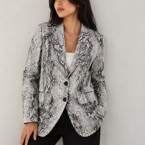 May include: A snakeskin-patterned blazer in shades of gray and black. The blazer has a notched collar, two black buttons, and two front pockets. The model is wearing black pants and a white top. The blazer is a stylish addition to any wardrobe.