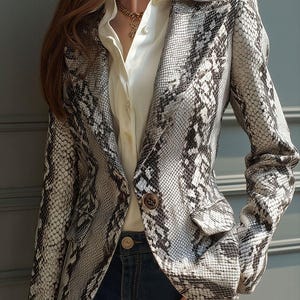 May include: A snakeskin-patterned blazer in shades of gray and white, featuring a notched lapel, two front pockets, and two brown buttons. The blazer is worn over a white collared shirt and dark blue jeans. A gold necklace is visible.