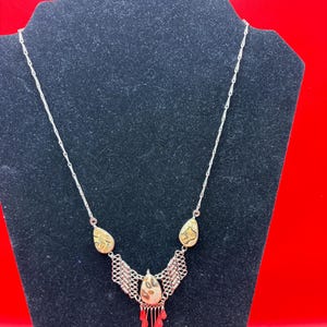 May include: A silver-toned necklace featuring a delicate chain and a central pendant arrangement. The design includes three teardrop-shaped stones with tan and brown mottled patterns, accented by intricate silver metalwork and dangling red drops.