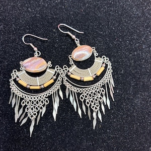 May include: A pair of silver-toned dangle earrings featuring an oval-shaped marbled stone at the top, a curved wire-wrapped section, horizontal light-colored cylindrical beads, and a bottom fringe of small silver-toned metal leaf-shaped charms.