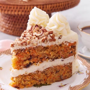 Può includere: Una fetta di torta di carote con due strati di pan di spagna punteggiato di arancione, separati e ricoperti da una spessa glassa bianca. La parte superiore è guarnita con noci pecan tritate e due ciuffi decorativi di glassa in cima.