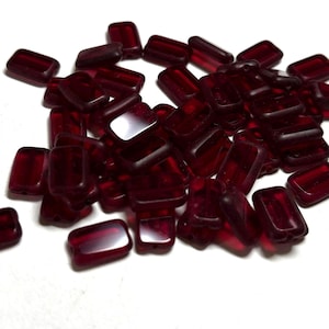 May include: A collection of small, rectangular, deep red translucent beads. Each bead features a smooth, polished surface with rounded corners and a flat profile, creating a uniform appearance across the scattered pile of individual pieces.
