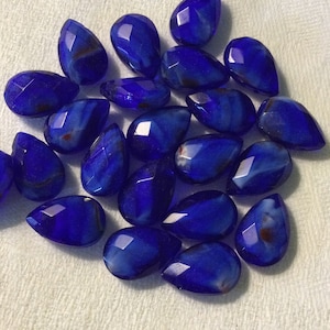 May include: A collection of faceted, teardrop-shaped blue gemstones. The stones have a deep, rich blue color with subtle variations and internal patterns. The gemstones are arranged on a white, textured surface.