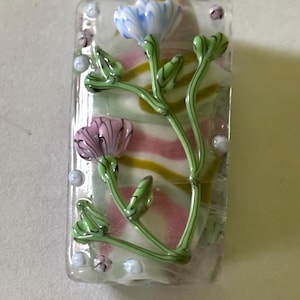 May include: A rectangular glass pendant with a floral design. The pendant features a light blue and pink flower with green stems and leaves. The background has a striped pattern of yellow, pink, and white. Small, clear beads are scattered around the edges.