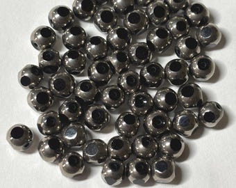 May include: A collection of small, spherical, metallic-toned beads with a polished, reflective surface. Each bead features a central hole, creating a uniform appearance across the group. The beads are arranged in a loose, scattered cluster.