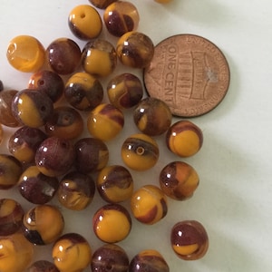 May include: A pile of brown and yellow glass beads. The beads are round and have a swirl pattern. There is a US penny in the image for scale.
