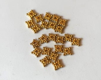 May include: A scattering of small, gold-coloured, square-shaped embellishments. Each piece has a four-point design with a central hole and small, rounded details. The embellishments are on a white surface.