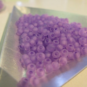 May include: A close-up of a pile of small, light purple beads. The beads are round and have a slightly frosted finish. They are arranged in a loose pile on a silver tray.