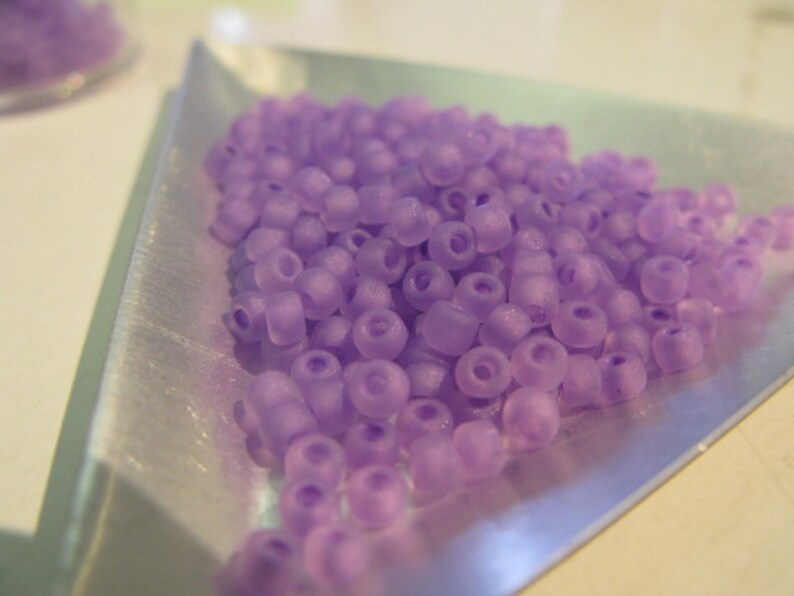 May include: A close-up of a pile of small, light purple beads. The beads are round and have a slightly frosted finish. They are arranged in a loose pile on a silver tray.