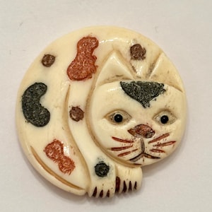 May include: A carved, cream-colored cat figurine with brown and black spots. The cat is curled up, with its head and paws visible. The eyes are black, and the nose and whiskers are detailed with brown lines.