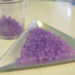 May include: A pile of small, light purple glass beads in a triangular silver tray. The beads are translucent and have a slightly frosted finish.