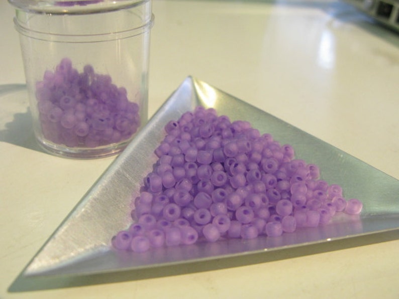 May include: A pile of small, light purple glass beads in a triangular silver tray. The beads are translucent and have a slightly frosted finish.