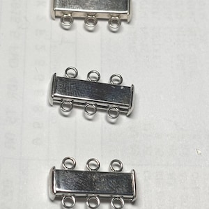 May include: Three silver-colored, rectangular jewelry clasps. Each clasp has three loops on each side for attaching to jewelry. The clasps are made of a shiny, metallic material.