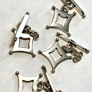 May include: Silver-toned cufflinks with a geometric diamond-shaped design. Each cufflink features a square center and four small ball accents at the corners. The cufflinks are connected by a chain and a bar.