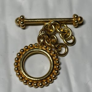 May include: A gold-toned toggle clasp for jewelry, featuring a circular ring with a beaded outer edge and a matching T-bar component. The two pieces are connected by a short, thick gold-toned chain link, creating a functional jewelry fastener.