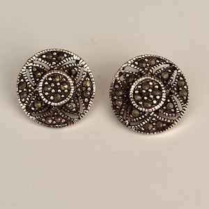 May include: A pair of silver earrings with a round, intricate design featuring a central cluster of marcasite stones.