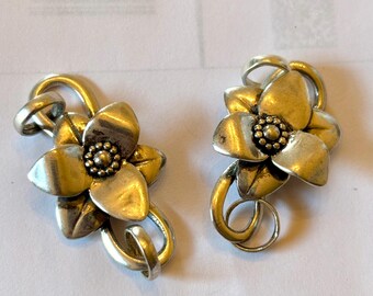 May include: A pair of silver-toned flower-shaped earrings. Each earring features a detailed floral design with five petals and a central cluster of small beads. The earrings have a curved, looped attachment.