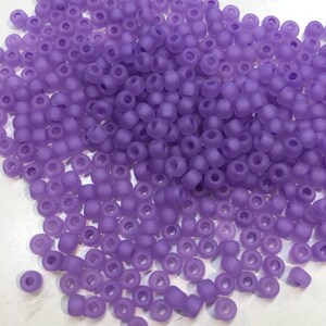May include: A large pile of small, round, opaque purple beads. The beads have a frosted finish and are scattered on a white surface.