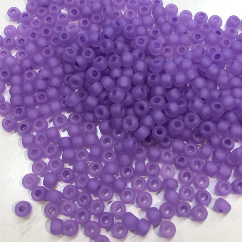 May include: A large pile of small, round, opaque purple beads. The beads have a frosted finish and are scattered on a white surface.