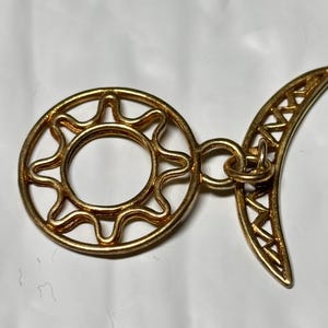 May include: A gold-toned metal pendant featuring a circular sun motif with wavy rays connected by a small jump ring to a crescent moon shape. The crescent moon has a decorative cutout pattern along its inner curve, creating a celestial design.