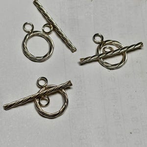 May include: Three silver-toned toggle clasps. Each clasp features a twisted, cylindrical bar and a circular ring with a smaller loop attached. The clasps are designed for jewelry making and crafting.