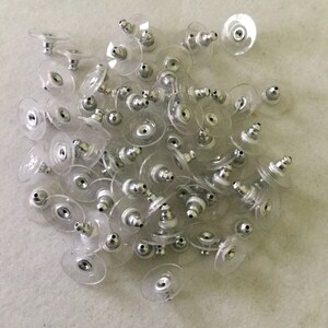 May include: A collection of numerous earring backs, each featuring a small metallic silver-toned post component securely embedded within a circular, flexible, and transparent plastic disc designed to provide support and comfort for earrings.