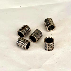 May include: Five silver-colored, cylindrical beads with a grid-like pattern. Each bead has a dark interior and a slightly tarnished appearance, suggesting an aged or vintage aesthetic. The beads are scattered on a white surface.