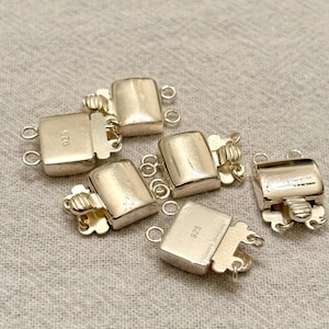 May include: A collection of silver-colored jewelry clasps. The clasps are rectangular with a rounded top and feature a small loop on each end for attachment. The clasps are marked with "925". The background is a neutral, textured fabric.