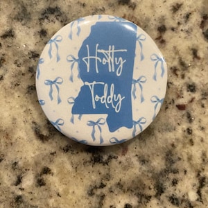 May include: A round button with a white background and a pattern of light blue bows. The center features a light blue outline of a state with the words "Hotty Toddy" in script.