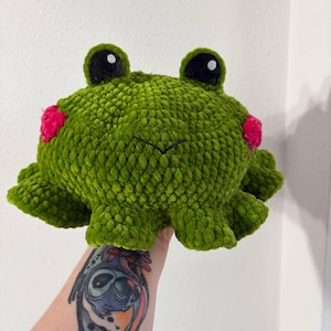 Könnte beinhalten: Ein handgemachter, grüner Plüschfrosch mit großen schwarzen Augen und rosa Akzenten. Der Frosch hat ein strukturiertes, flauschiges Aussehen und einen lächelnden Mund. Der Körper des Frosches ist rund mit gerüschten Kanten.