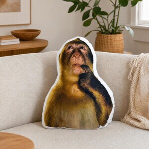 May include: A decorative pillow shaped like a monkey, featuring a realistic image of a monkey with its hand to its chin. The pillow has a white background and is placed on a beige sofa. The monkey's fur is brown and tan.