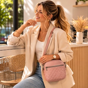 May include: A light pink quilted crossbody bag with a zipper closure and a long strap. The bag is worn over the shoulder. The person is wearing a cream-colored blazer, a white top, and blue jeans. A gold watch and jewelry are also visible.