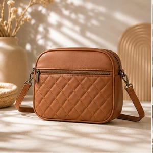 Pode incluir: Uma bolsa transversal acolchoada marrom com formato retangular. A bolsa possui um bolso frontal com zíper e uma alça longa e ajustável. A bolsa é feita de um material liso e possui um padrão de losangos.