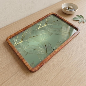 Puede incluir: Bandeja rectangular con fondo verde y diseño de hojas doradas. La bandeja tiene un borde marrón texturizado. Un cuenco pequeño con piedras y una hoja están en el fondo.