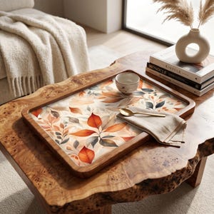 Puede incluir: Una bandeja de servicio rectangular de madera con un patrón impreso de hojas de otoño naranjas, marrones y grises. La bandeja contiene un pequeño cuenco de cerámica, una servilleta de lino beige doblada y una cuchara dorada sobre una mesa de madera.