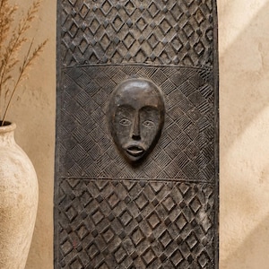 May include: A tall, dark brown wooden shield with a rounded top and bottom. The surface features intricate hand-carved geometric patterns, including cross-hatching and diamond shapes, with a central carved human face in relief at the center.