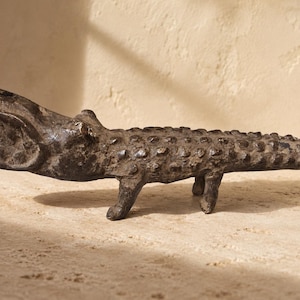 Op de afbeelding: Een donkerbruin, getextureerd metalen beeldje van een alligator met de bek open. Het wezen heeft een bobbelige, geschubde rug en vier korte poten, en staat in een horizontale positie met een lange, taps toelopende staart aan de rechterkant.