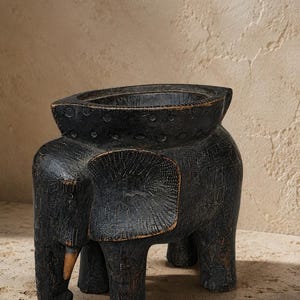 Peut inclure: Un tabouret en bois sculpté en forme d'éléphant, de couleur gris foncé. Le tabouret a un dessus en forme de bol et quatre pieds robustes. Les oreilles et la trompe de l'éléphant sont visibles, et le bois a une finition texturée et vieillie.