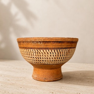Puede incluir: Un cuenco artesanal de color terracota con base de pedestal. El cuenco presenta un diseño decorativo calado en color crema en el centro, con líneas horizontales cerca del borde. El cuenco es probablemente vintage o antiguo.