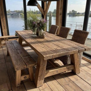 Op de afbeelding: Een rustieke houten eettafel met een bijpassende bank en rieten stoelen. De tafel en bank zijn gemaakt van verweerd hout, met natuurlijke nerven en knopen. De setting is in de buurt van een water, met grote ramen die een schilderachtig uitzicht bieden.