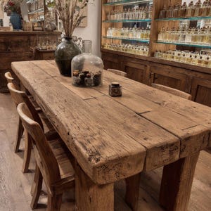 Peut inclure: Une grande table en bois rustique avec une finition naturelle, entour&eacute;e de chaises en bois. Des bouteilles et bocaux en verre d&eacute;coratifs sont plac&eacute;s sur la table. Des &eacute;tag&egrave;res remplies de flacons de parfum sont visibles en arri&egrave;re-plan.