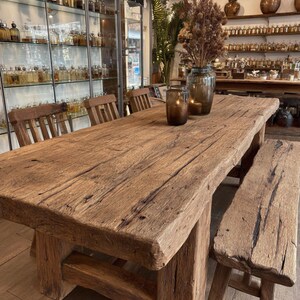 Peut inclure: Une table &agrave; manger rustique en bois avec un banc assorti. La table a une surface &eacute;paisse et vieillie et des pieds robustes. Un vase avec des fleurs s&eacute;ch&eacute;es et un bougeoir sont pos&eacute;s sur la table. Les chaises sont &eacute;galement en bois.