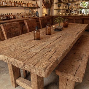 Peut inclure: Une grande table &agrave; manger en bois rustique avec un banc assorti. La table est en bois r&eacute;cup&eacute;r&eacute;, pr&eacute;sentant une texture vieillie et un grain naturel. Des bouteilles en verre ambr&eacute; et une petite plante d&eacute;corent la surface. Les chaises sont en cuir marron.