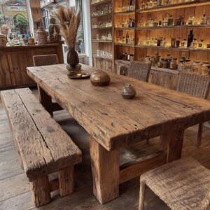 Peut inclure: Une grande table &agrave; manger en bois rustique avec un banc assorti et des chaises en osier. La table est faite de bois &eacute;pais et vieilli et a un aspect naturel et non fini. Des vases d&eacute;coratifs et un plateau avec des bougies sont pos&eacute;s sur la table. Des &eacute;tag&egrave;res remplies de bouteilles en verre sont en arri&egrave;re-plan.